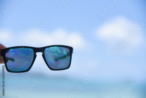 Man hand holding sunglasses on the beach with blurred sea and blue sky background. Summer day concept. Vacation holidays background wallpaper.