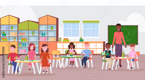 woman teacher teaching mix race boys and girls preschool modern kindergarten children classroom with chalkboard desks chairs kid room interior full length flat horizontal