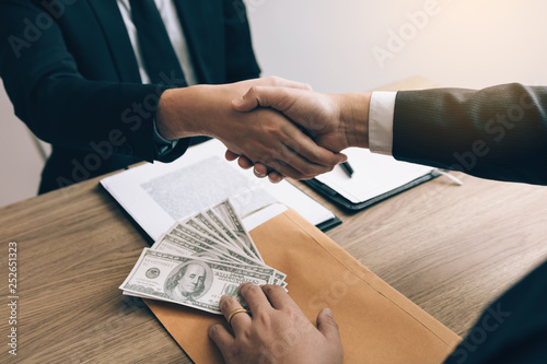 Canvas Print Two corporate businessmen shaking hands while one man places money on envelope document in office room with corruption concept