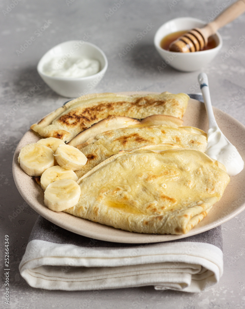 Thin pancakes or crepes with natural yogurt, honey and banana on a plate. Light concrete background. Copy space. Thin pancakes or crepes with natural yogurt, honey and banana on a plate. Light concrete background. Copy space.