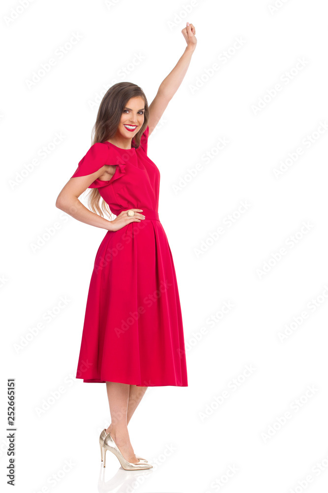 Woman In Elegant Red Dress And Gold High Heels Is Standing With Arm Raised, Looking At Camera And Smiling