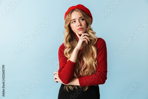 Portrait of stylish blond woman 20s wearing red beret looking aside while standing, isolated over blue background in studio photo