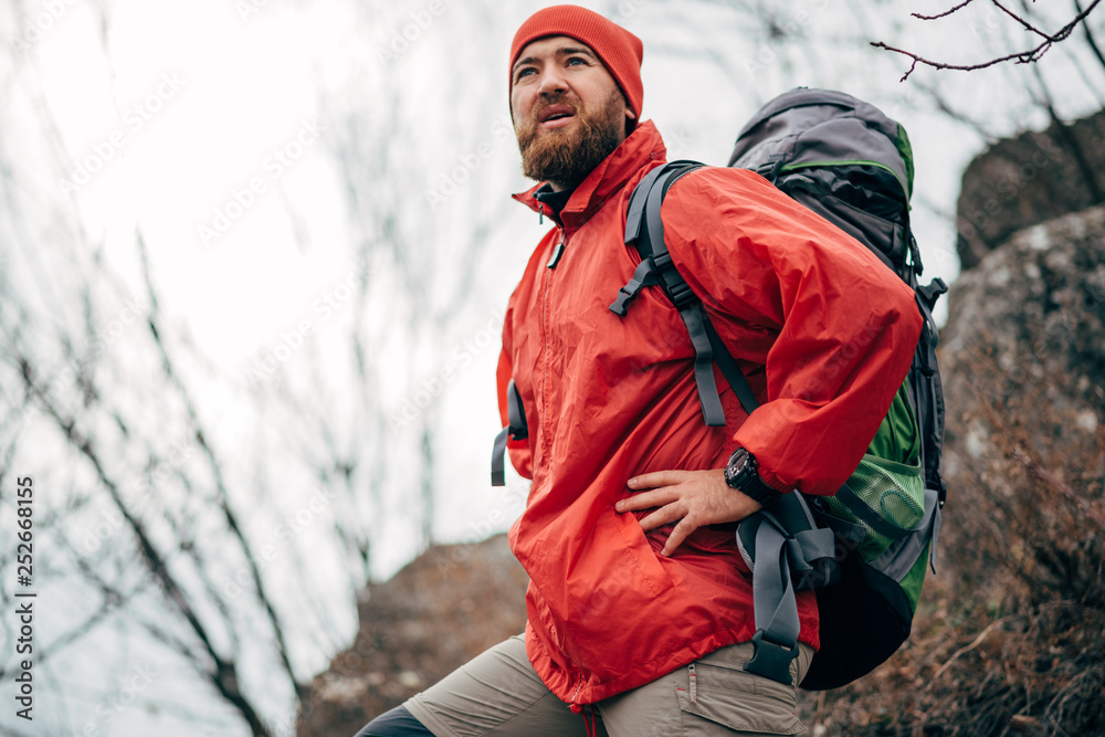 Portrait of young male hiking in mountains wearing red clothes exploring new place. Traveler bearded man trekking and mountaineering during his journey. Travel, people, sport and lifestyle concept