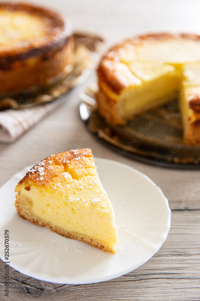 Ein Stück Käsekuchen oder Quarkkuchen auf weißem Teller dahinter ganzer und angeschnittener Kuchen auf Blechform und graues Holz als Untergrund Ein Stück Käsekuchen oder Quarkkuchen auf weißem Teller dahinter ganzer und angeschnittener Kuchen auf Blechform und graues Holz als Untergrund