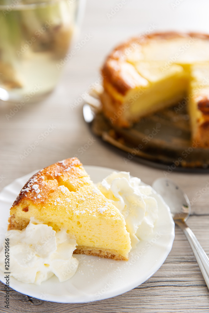 Ein Stück Käsekuchen oder Quarkkuchen auf weißem Teller dahinter ganzer und angeschnittener Kuchen auf Blechform und graues Holz als Untergrund Ein Stück Käsekuchen oder Quarkkuchen auf weißem Teller dahinter ganzer und angeschnittener Kuchen auf Blechform und graues Holz als Untergrund