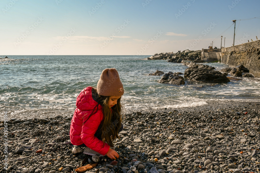 A little girl playing with the pebbles next to the water's edge of a beach in a sunny winter day, Boccadasse, Genoa, Liguria, Italy