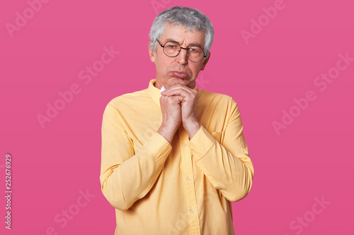 Studio portrait of old grey haired male. Offended man with sad look in yellow shirt and on pink background. Seniour with pouty lips, someone hurts elderly man. Pensioner and people concept. photo