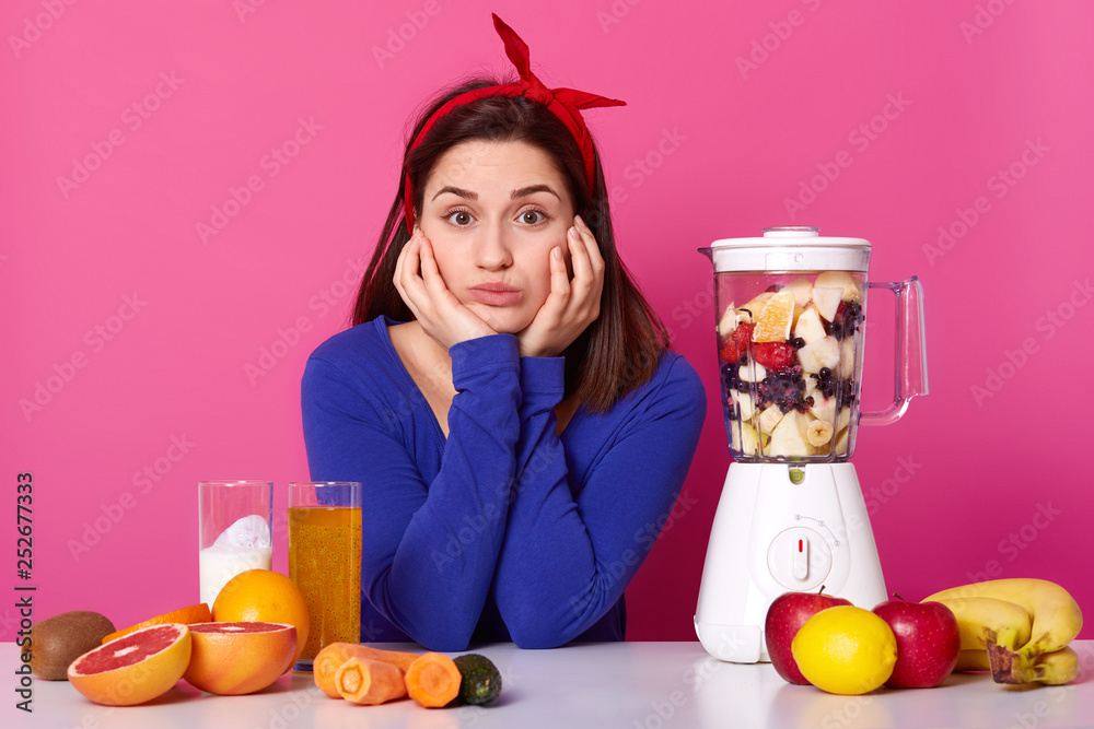 Sad woman sits at white table with blender filled with fresh fruit, makes smoothie for breakfast, touches cheeks and holds chin, wears blue jumper, isolated over pink background, being vegetarian Sad woman sits at white table with blender filled with fresh fruit, makes smoothie for breakfast, touches cheeks and holds chin, wears blue jumper, isolated over pink background, being vegetarian