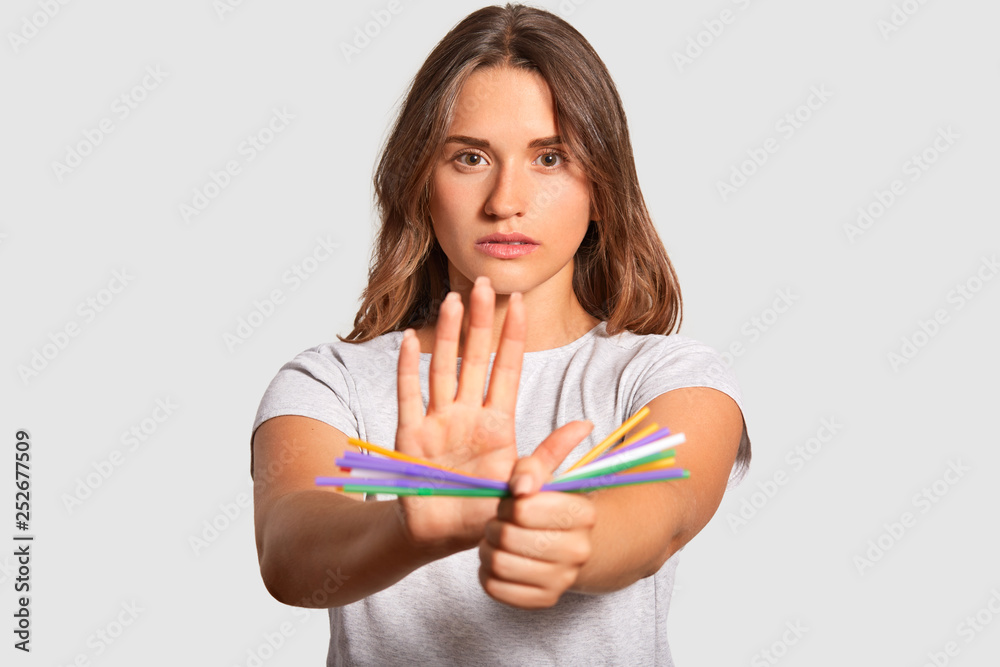 Horizontal shot of serious European young woman holds colourful plastic straws in both hands, demonstrates her protest of plastic pollution, wears casual clothes, stands indoor. Ecology concept Horizontal shot of serious European young woman holds colourful plastic straws in both hands, demonstrates her protest of plastic pollution, wears casual clothes, stands indoor. Ecology concept