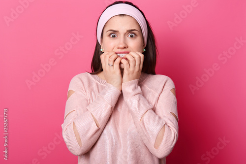 Woman with both hands in front of mouth. Scared girl in pink sweater and hairband, isolated over rose background. Lady wathes horror film alone. Female afraids of something. People concept. photo