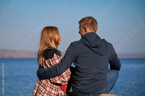 Young beautiful European couple is sitting on the big stone against picturesque lake, they looking to each other with tenderness.