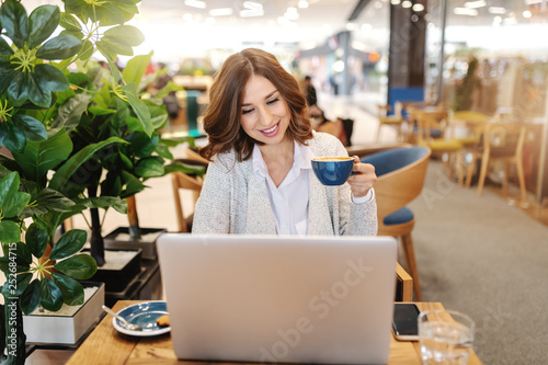 Charming brunette with toothy smile using laptop and drinking coffee in cafeteria.