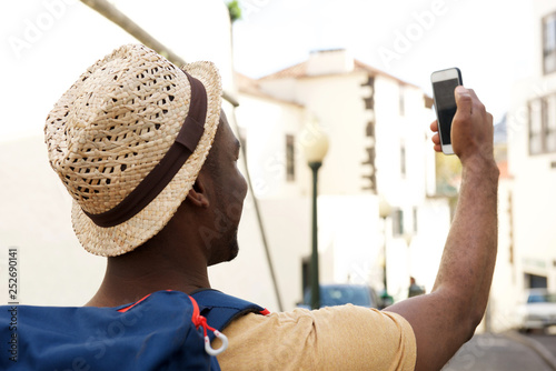 behind of african american male tourist with bag and hat taking selfie