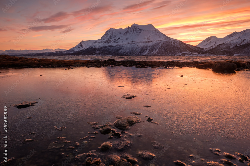 Frozen pool in foreground with beautiful reflection of snowcapped mountain in background at cloudy and colorful sunrise, Kvalöya, Norway