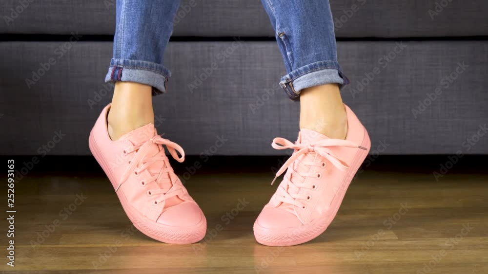 Woman is sitting on gray sofa and shows her legs in comfortable coral sneakers on wooden floor Woman is sitting on gray sofa and shows her legs in comfortable coral sneakers on wooden floor