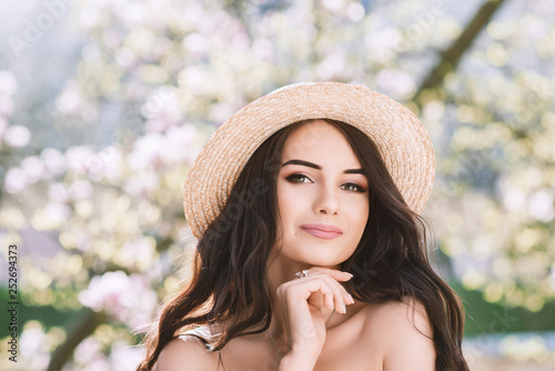 Outdoor close up portrait of young beautiful happy smiling lady with long dark hair, makeup, wearing straw hat, posing near blooming tree. Copy, empty space for text