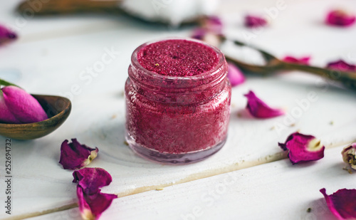 Homemade natural lip scrub from jojoba oil, cherry, rose oil and cane sugar in a small plastic jar. Scrub stands on a wooden table, around the rose petals. Handmade cosmetics, side view
