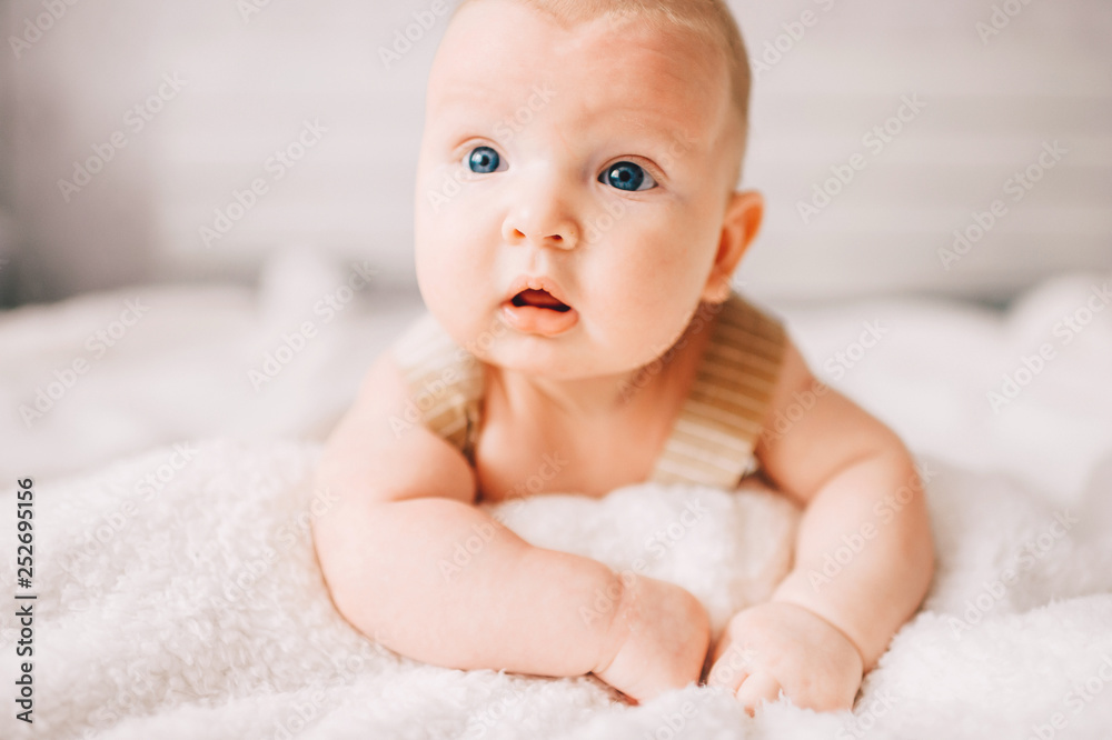 Adorable lovely newborn male baby boy with smiling emotional happy face lifestyle indoor portrait. Funny infant child lying on stomach on bed with white wall on background. Carefree childhood. Adorable lovely newborn male baby boy with smiling emotional happy face lifestyle indoor portrait. Funny infant child lying on stomach on bed with white wall on background. Carefree childhood.