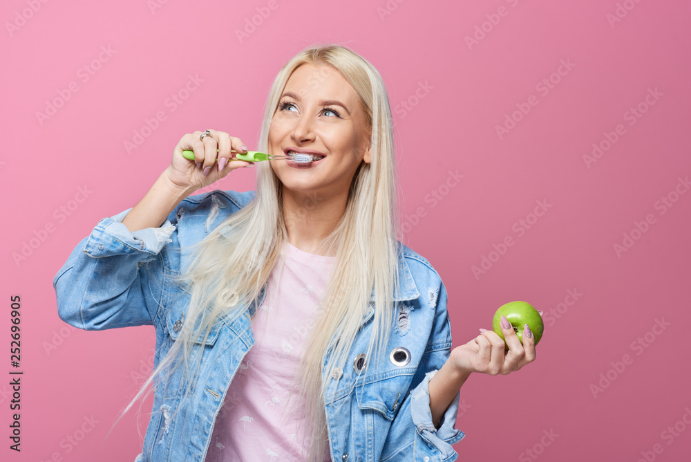 Φωτογραφία Close up portrait of young blond woman with braces on teeth holding green apple and toothbrush