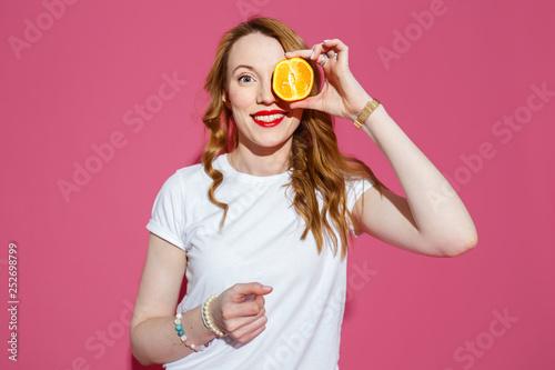 Beautiful blonde holding fresh orange, posing, smiling on pink background in studio. Bright colors, hard light.