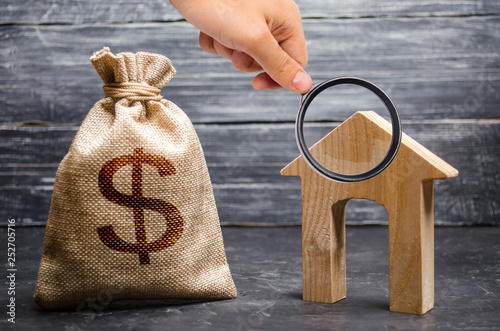 Magnifying glass is looking at a bag with money and a house with a large doorway. Concept of real estate acquisition and investment. Affordable cheap loan, mortgage. Taxes, income. Building houses