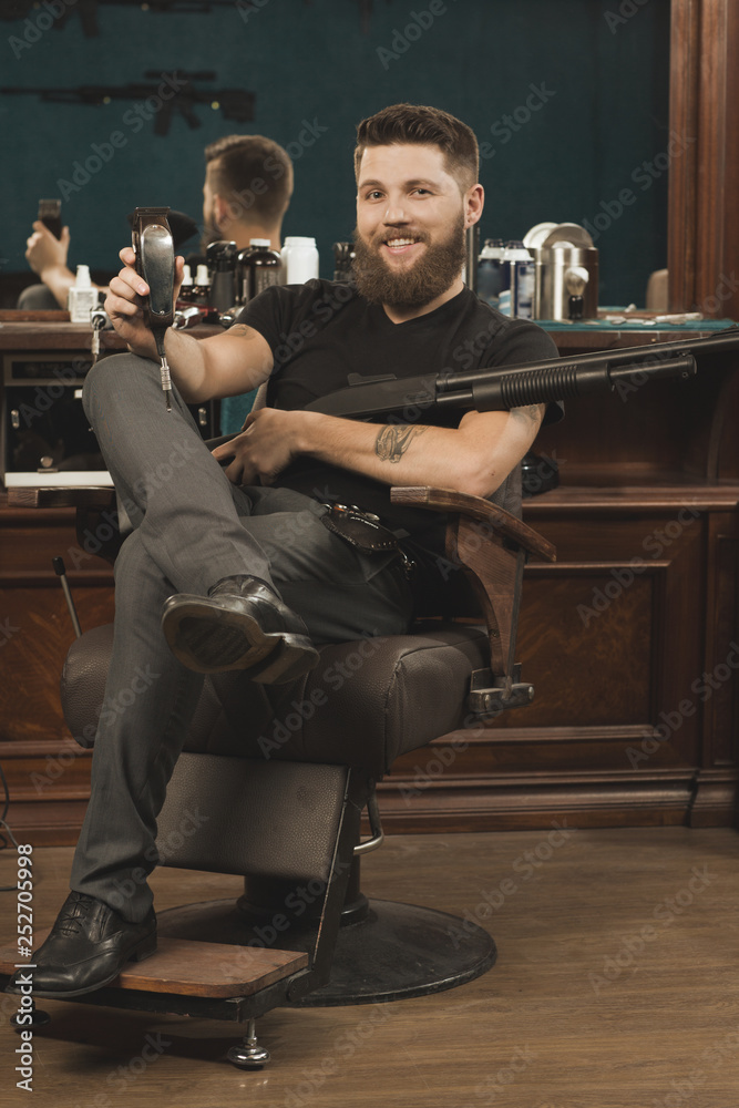 Obraz premium You will love my styling! Handsome tough and manly bearded barber sitting in a chair with a trimmer in his hand and holding a Winchester gun and smiling happily