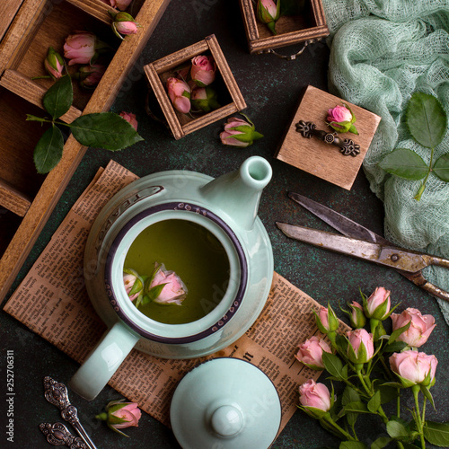 Overhead shot of composition with green teapot with tea, little wooden chest of drawers, pink roses, green gauze and old scissors on dark green concrete background photo