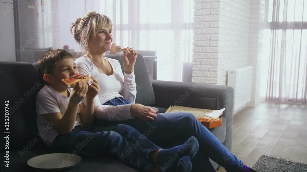 Cute preadolescent son with adult mother eating pizza and looking at TV screen showing funny child cartoon. Cheerful family having fun sitting on sofa in living room watching comic movie on TV Cute preadolescent son with adult mother eating pizza and looking at TV screen showing funny child cartoon. Cheerful family having fun sitting on sofa in living room watching comic movie on TV