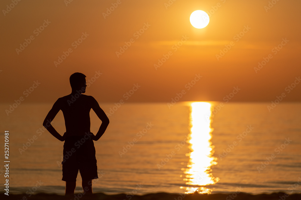 dark silhoutte of unrecognizable man standing on beach in evening summer in front of sunset with sun light falling on water.  Beautiful nature  seascape. Sun light reflected in ocean.
