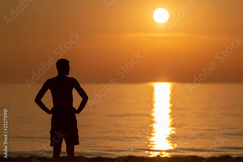 dark silhoutte of unrecognizable man standing on beach in evening summer in front of sunset with sun light falling on water. Beautiful nature seascape. Sun light reflected in ocean.