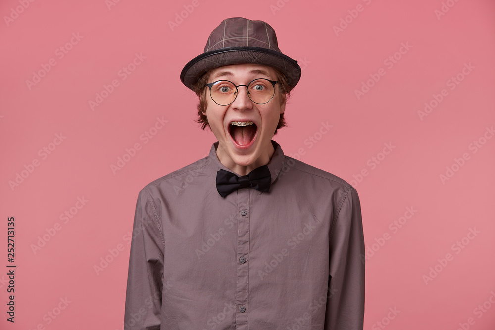 Guy opened his mouth in surprise, is overwhelmed with positive emotions happiness joy doesn't believe in his success, luck, dressed in shirt hat and black bowtie glasses has brackets isolated on pink