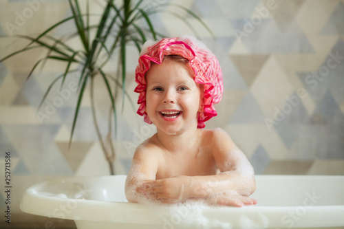 A little girl 3-4 years old in a pink cap, taking a bubble in a beautiful bathroom with a light background. Children's hygiene. Shampoo, hair treatment and soap for children. White bubble bath