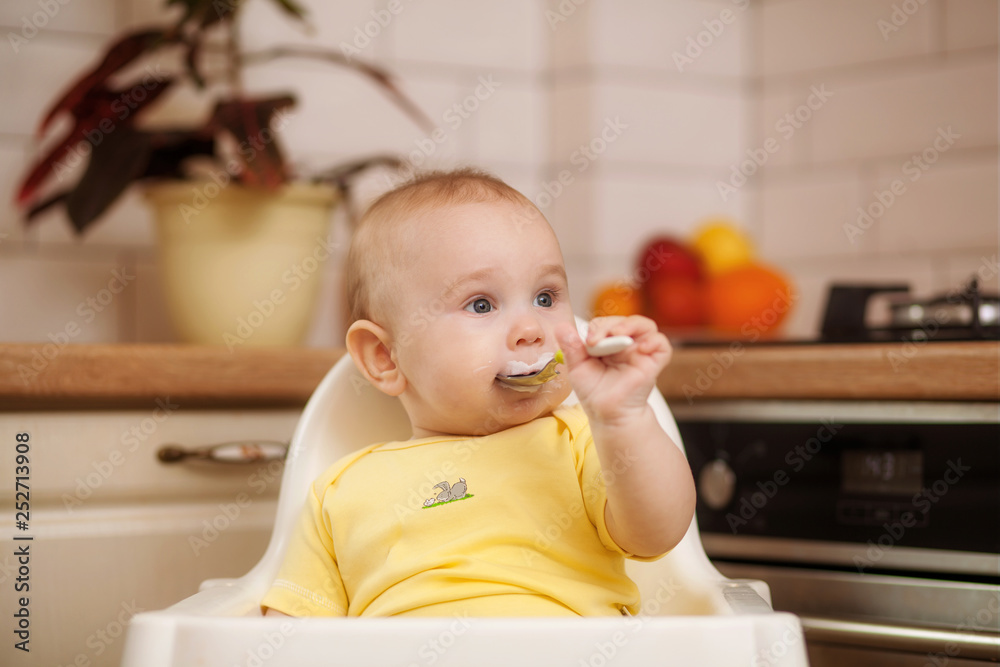 Fototapeta premium child up to a year sitting with a spoon in a chair for feeding in the kitchen