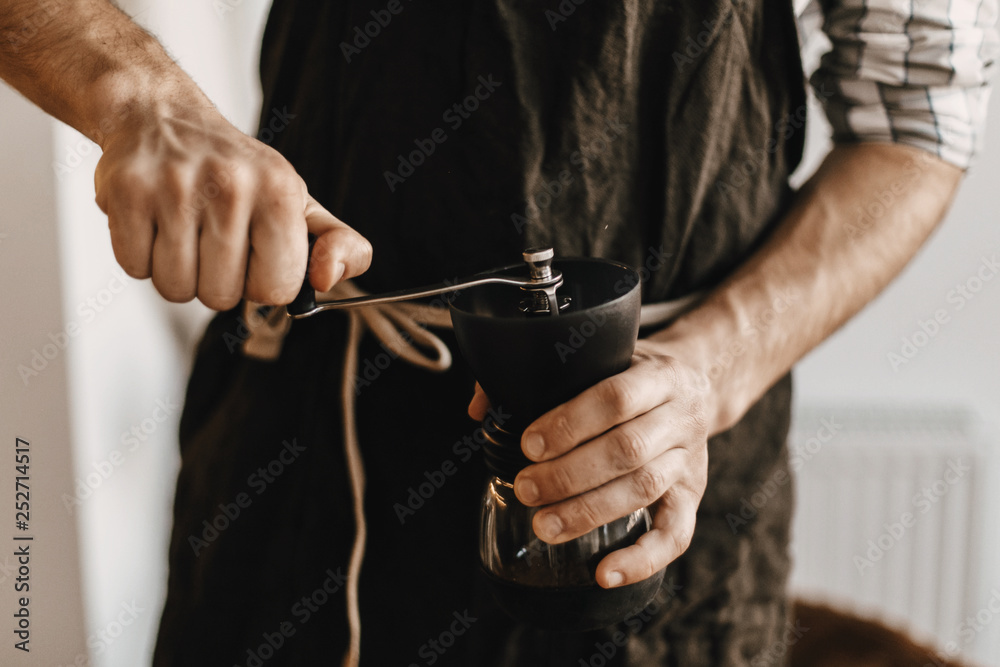 Professional barista in black stylish apron grinding coffee for aeropress, alternative brewing method. Hands holding manual grinder  with coffee beans. Items for an alternative coffee