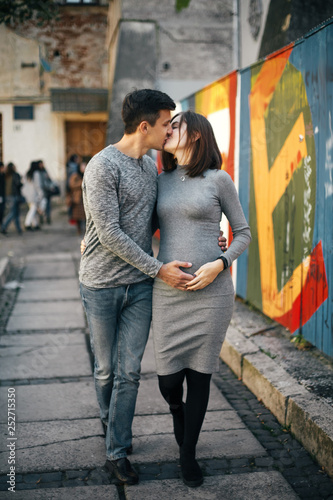 Stylish pregnant couple holding hands on baby bump and walking in city street. Happy young parents, mom and dad, hugging belly and smiling. Parenthood concept. True happiness