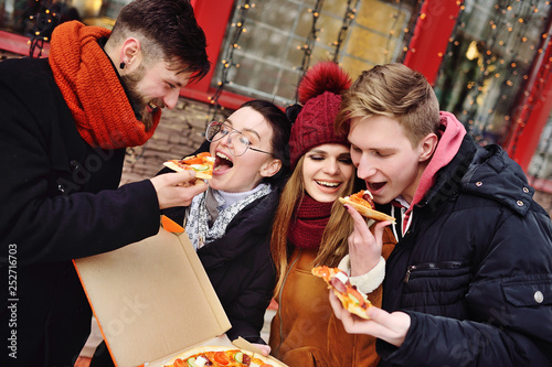 group of friends with a box of pizza smiling and eating pizza on the street