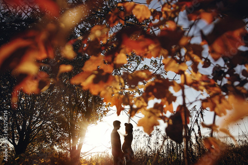 Stylish pregnant couple holding hands on belly in sunny light in autumn park among leaves. Happy young parents, mom and dad, hugging baby bump, enjoying beautiful moment at sunset. Creative photo