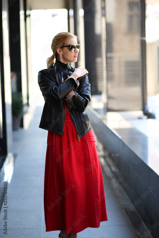Sexy Beauty Blond hair Girl with red Lips wearing red long dress and leaf jacket is posing outdoor at urban area of the city. Luxury Woman in stylish glasses and black leaf jacket.