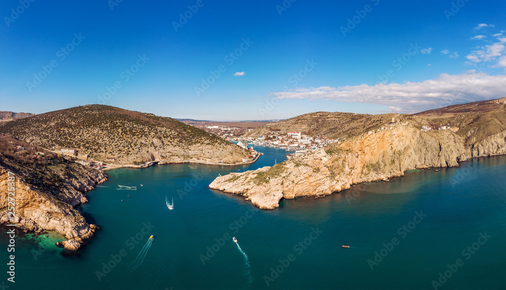 Aerial panoramic view of Balaklava bay in Crimea, mountain cliffs and sea with ships. Beautiful nature panorama landscape, town among hills and black sea coast from air, drone shot