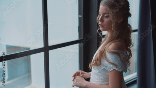 sweet lady with blond hair and tnder skin posing for camera in medieval room, long white dress with open soulders, cute curls of blond hair in young girl, maid with tender nude makeup video portrait photo