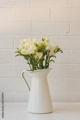 Fototapeta Naklejka Na Ścianę i Meble -  Vertical view of white freesias in jug on table against painted brick wall (selective focus)