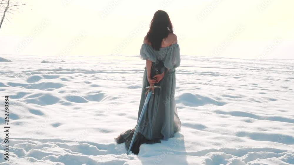 cute brunette walks along snowy field, a girl with long dark hair has her back to the camera, no face on video, lady in vintage gray adorable light dress pulls royal weapon of snow behind her