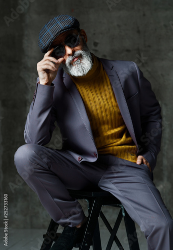 Brutal senior white bearded man in classic grey suit, green sweater, cap and aviator sunglasses sitting on a hight stool stylish fashionable men  photo