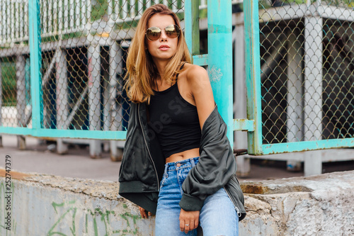 Fashion portrait of trendy young woman wearing sunglasses, jeans with halls and bomber jacket in the city