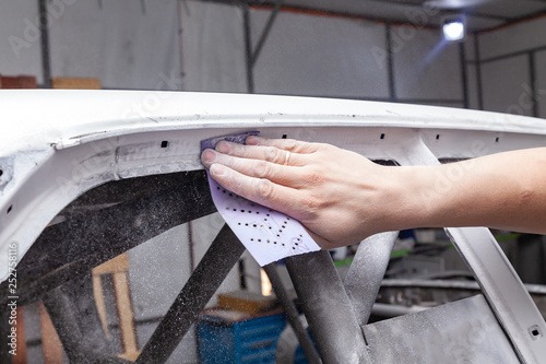 The body repairman grinds the white car's frame with purple emery paper in preparation for painting after applying putty in a vehicle repair workshop and auto service, dust crumbles down. photo