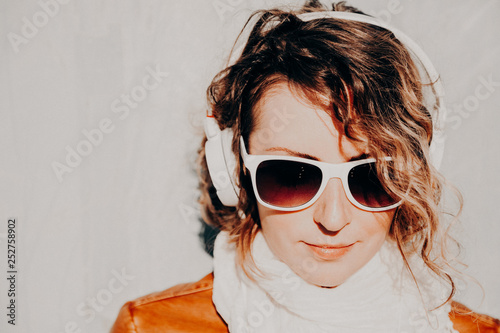 Beautiful young woman in a sunglasses and headphones listening to music standing at the wall. Casual style concept
