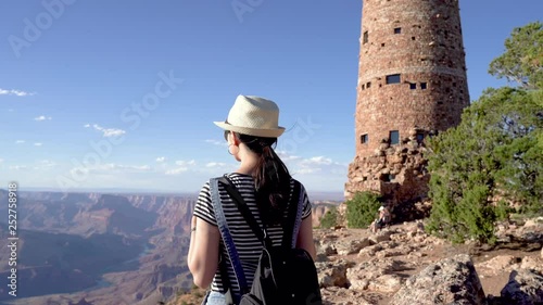 asian woman hiker with backpack cheerfully waving hand saying hi to camera. young college girl love traveling visiting famous attraction grand canyon standing next to old watch tower made by brick photo