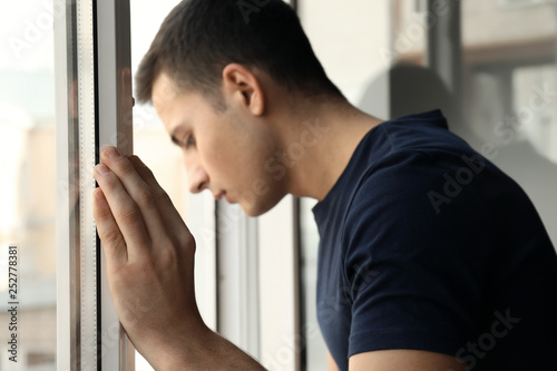 Depressed young man standing near window and thinking about suicide