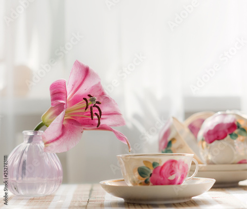 The tea cup is decorated with nicely pink flowers. Next to the vase is a vivid living flower of a similar color.