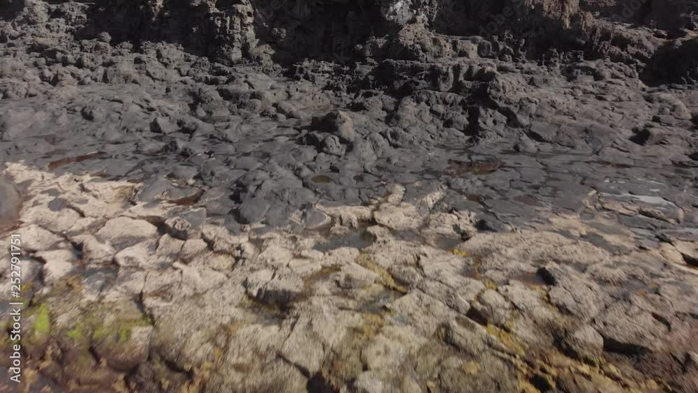 Aerial shot. Volcanic surface of the ocean coastline. Close-up of dark gray and yellow stones. Dolly shot from the turquoise color of the ocean waves. Aerial shot. Volcanic surface of the ocean coastline. Close-up of dark gray and yellow stones. Dolly shot from the turquoise color of the ocean waves.